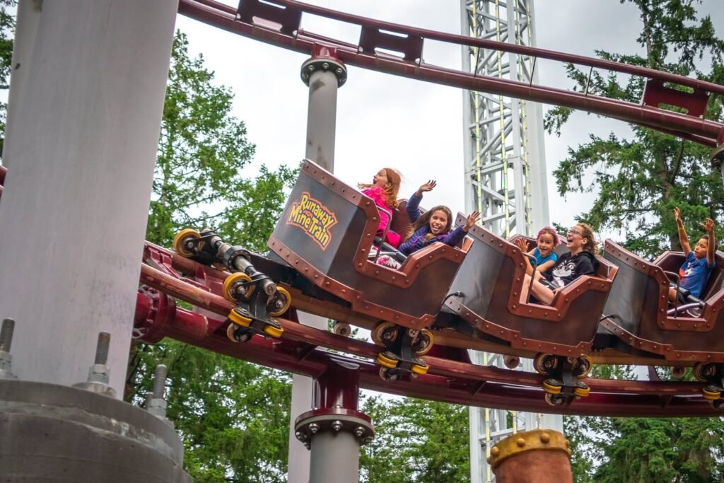 Riders on an outdoor roller coaster with raised hands, surrounded by green trees under a cloudy sky.
