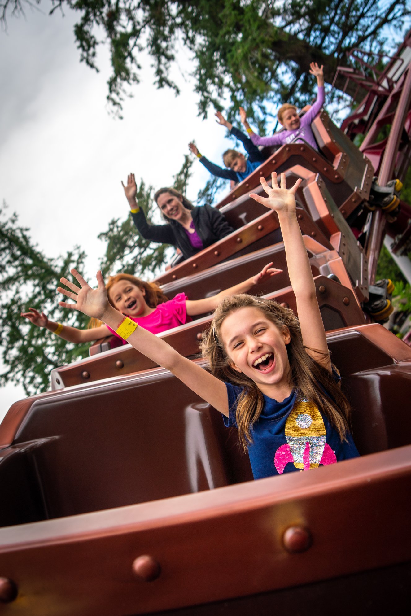Group of excited people with raised hands enjoy a roller coaster ride outdoors.
