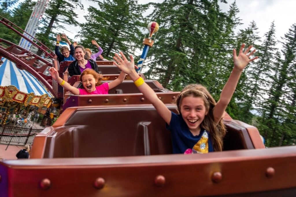 A group of people ride a roller coaster with raised hands, smiling eagerly, surrounded by green trees.
