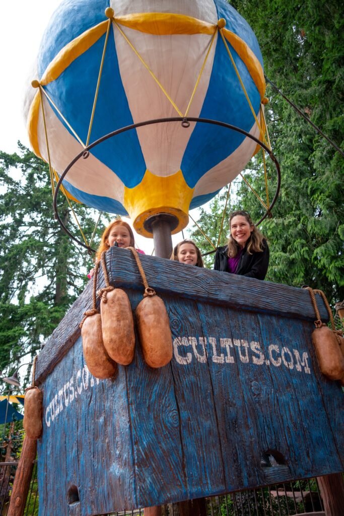 Three women smiling from inside a colorful hot air balloon sculpture. The balloon feature is vibrant with blue, pink, and orange sections.