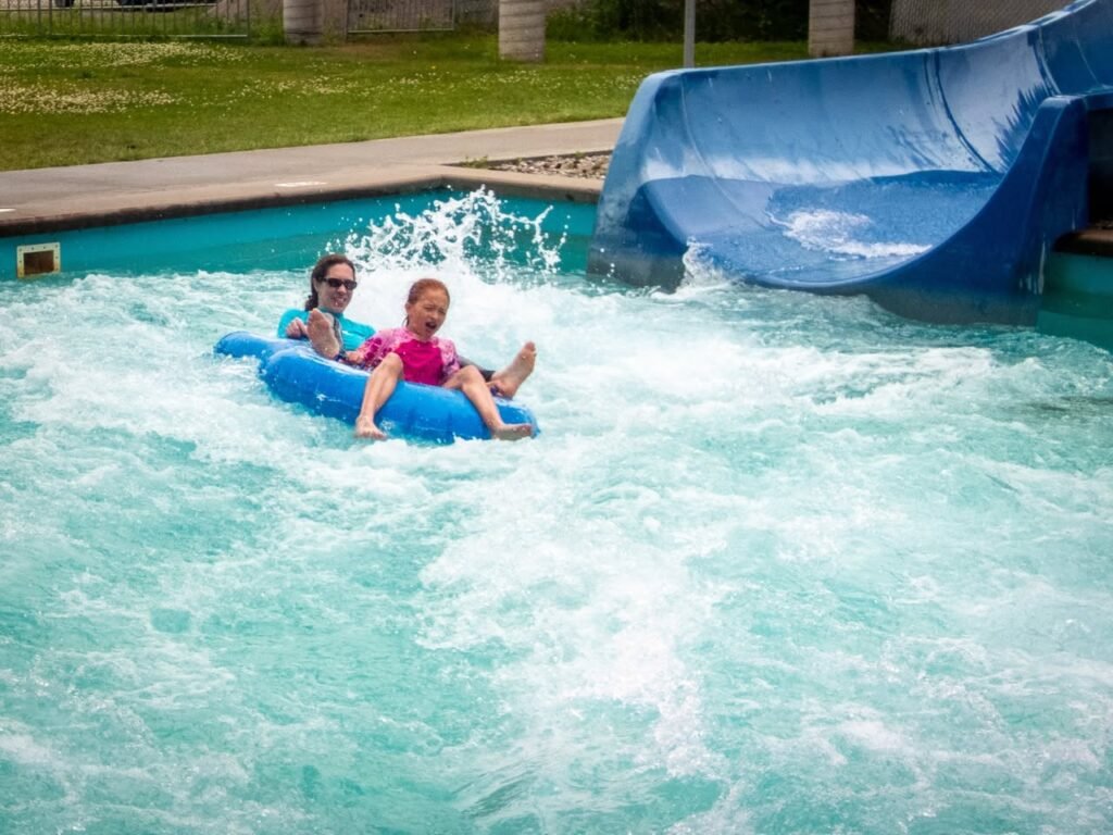 Two people on a blue inner tube sliding into a wave pool. The water splashes around them next to the blue water slide structure.