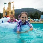 Two children enjoy a splash in a pool with castle-like structures and mountains in the background.