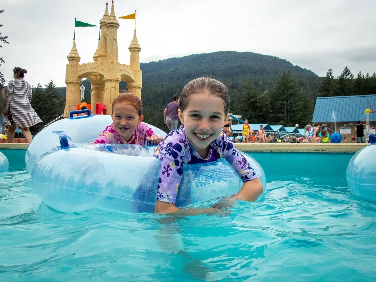 Two children enjoy a splash in a pool with castle-like structures and mountains in the background.