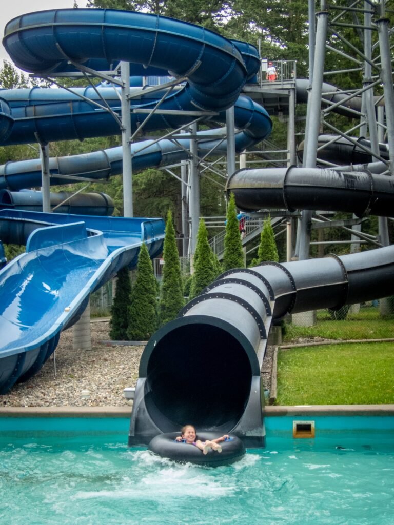 A visitor emerges from a black water slide tube into a turquoise pool. Multiple blue and black water slides surround the water park area.