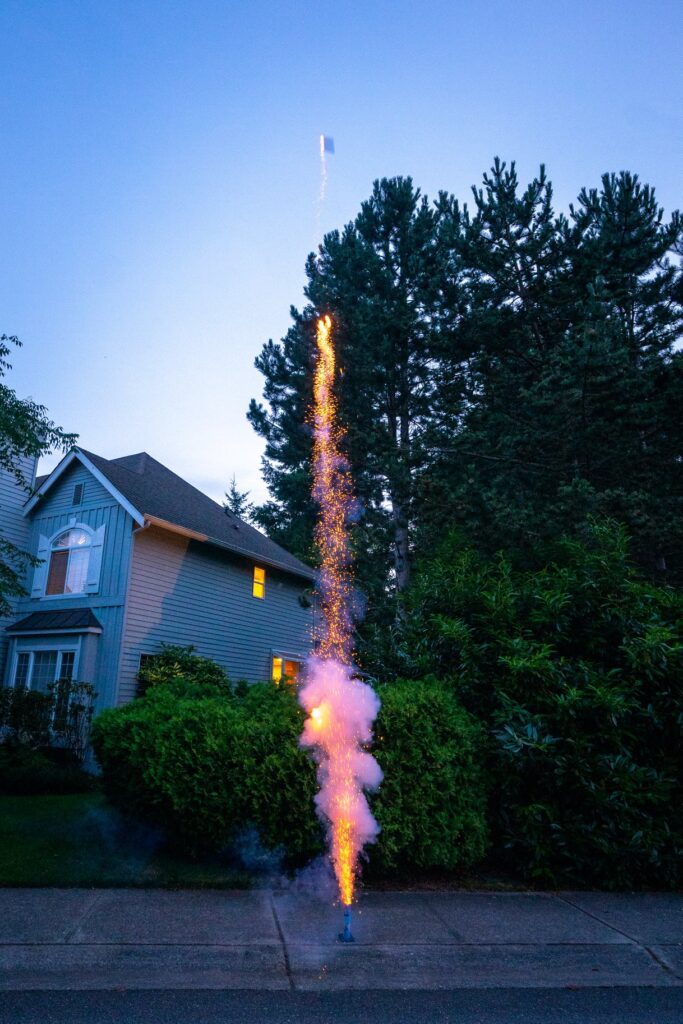 A vertical firework illuminates the night sky with orange and pink hues beside a suburban home and pine trees.