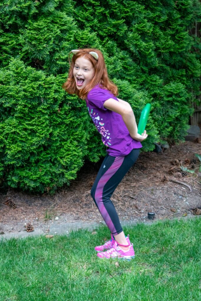 A playful child wearing a purple shirt and cat ears hits a green water gun to the rear while laughing outdoors.