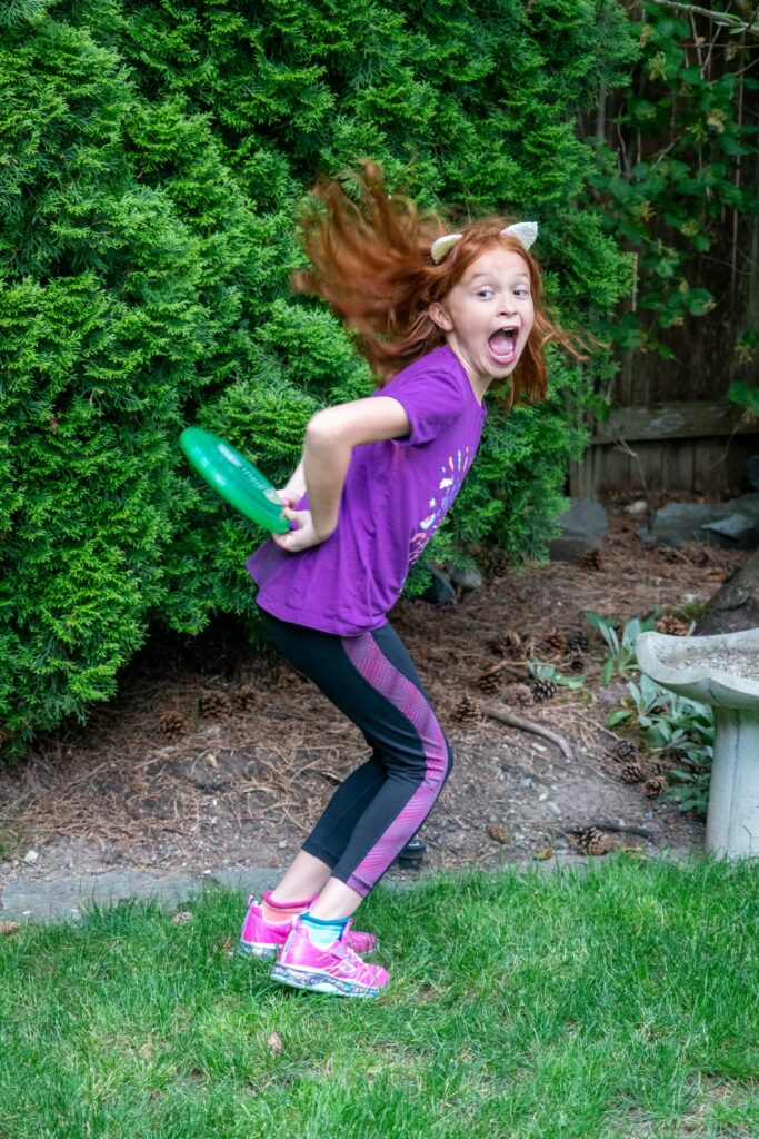 A child leaping in a backyard garden with a green disc. Lush foliage frames the joyful outdoor moment.