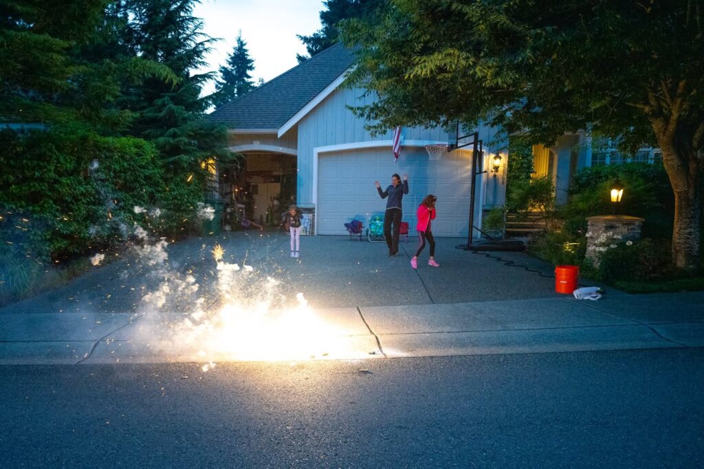 A bright firework burst illuminates a driveway in front of a house with a garage. Lush trees and warm exterior lights frame the evening scene.