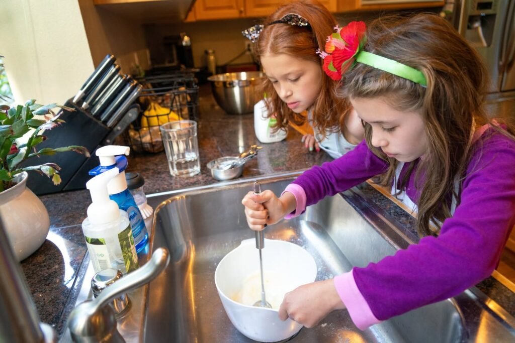 Two young girls mixing ingredients in a white bowl at a kitchen sink. A close-up of hands holding a whisk.