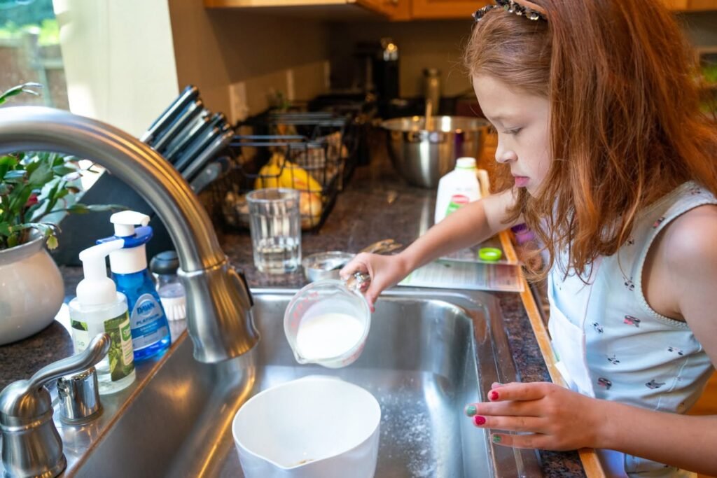 A child pours ingredients into a measuring cup at a kitchen sink, surrounded by household items and a nearby plant.