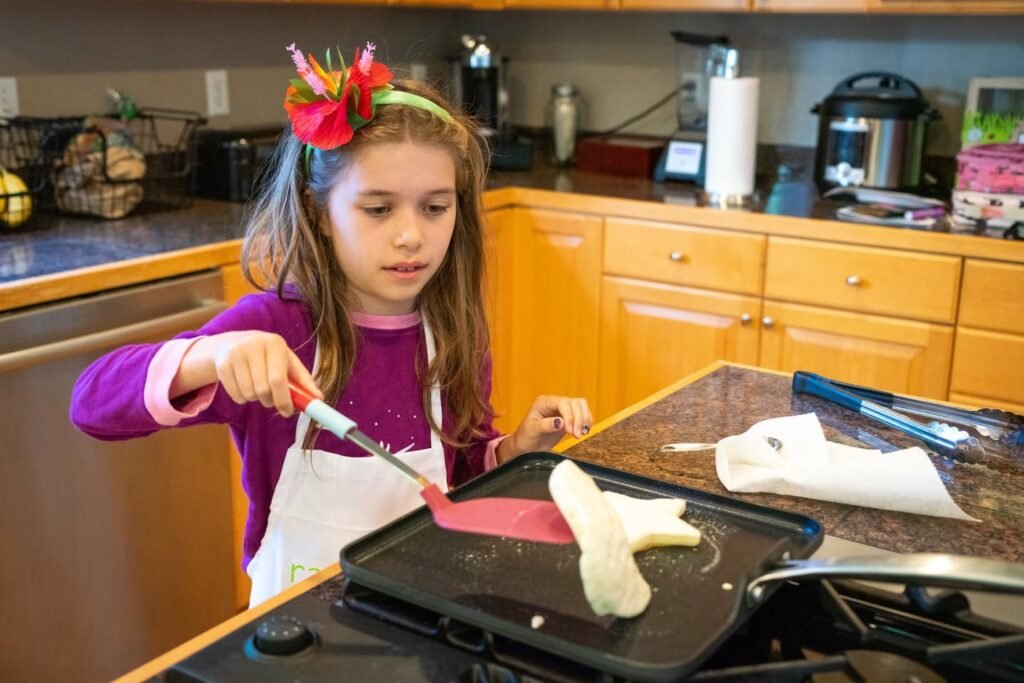 A child wearing a white apron stands at a kitchen counter working with a sausage on a griddle. The child uses a spatula to handle the food.