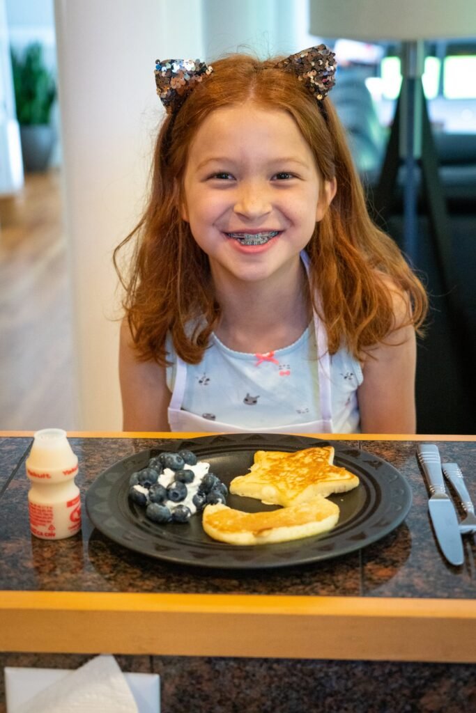 A young child with red hair and a hair clip smiles at a table with a plate of pancakes, blueberries, and a small bottle of yogurt.