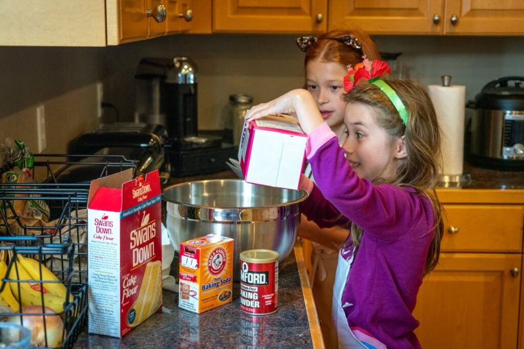 Two children prepare food together in a home kitchen. One pours ingredients into a mixing bowl as baking supplies sit on the counter nearby.