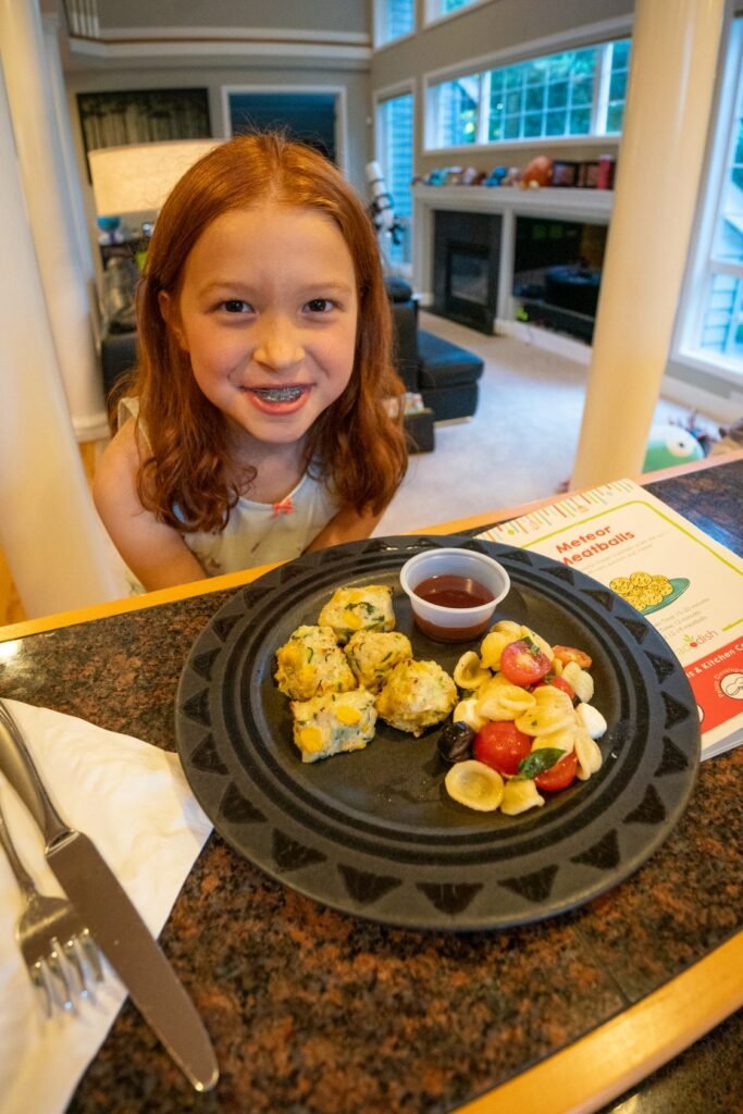 A smiling young girl looks on from behind a table with a black plate of diced food items.