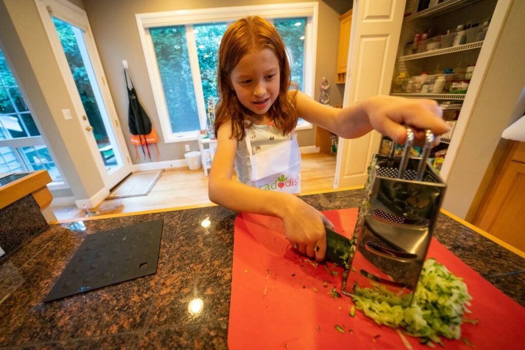 A child using a box grater on a red cloth over a granite countertop in a bright kitchen.