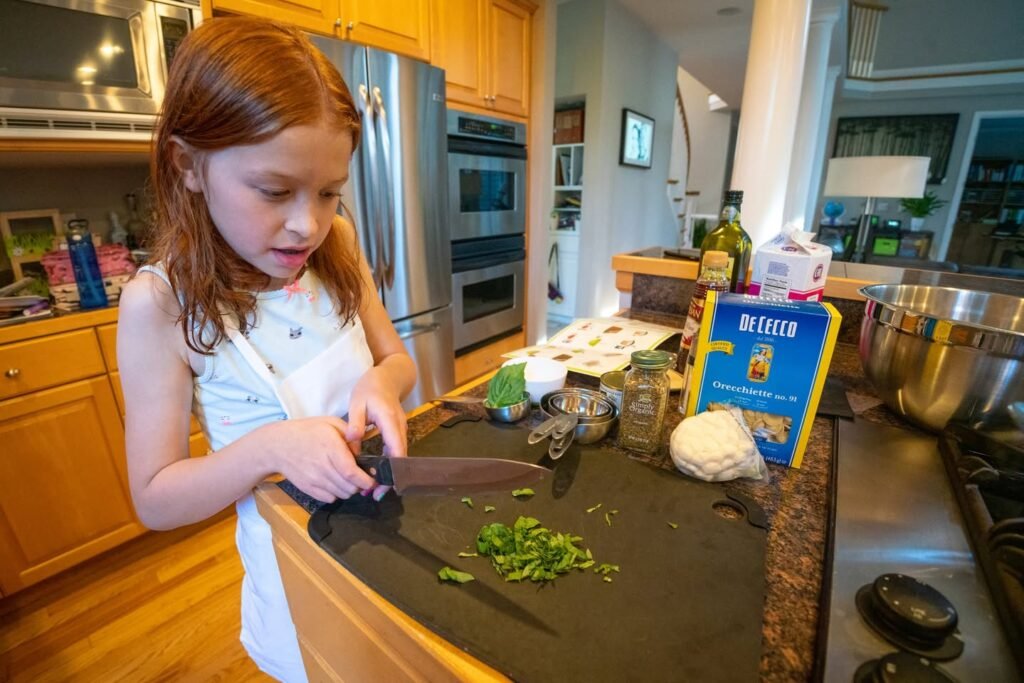 A young person chopping fresh green herbs on a cutting board in a home kitchen. A blue box of baking soda is visible on the counter next to a stainless steel oven and refrigerator.