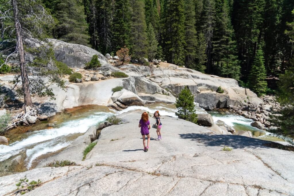 Two people exploring a rocky trail beside a rushing mountain stream surrounded by dense evergreen forest.