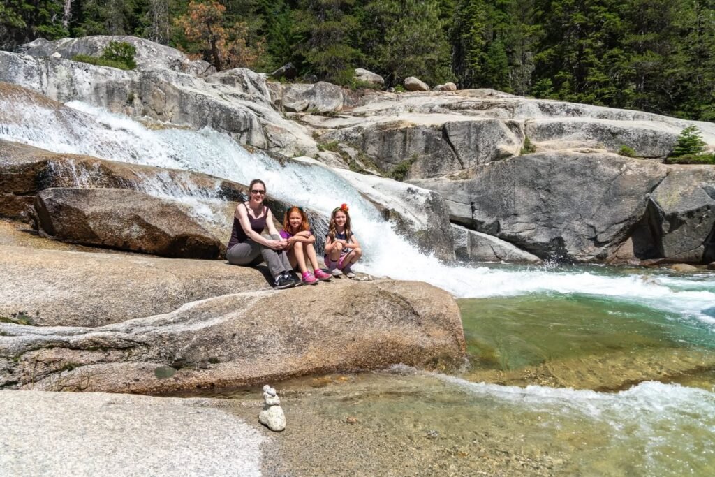 A family relaxes on a large flat rock beside a cascading waterfall in a lush forest setting. The clear water flows over smooth granite formations.