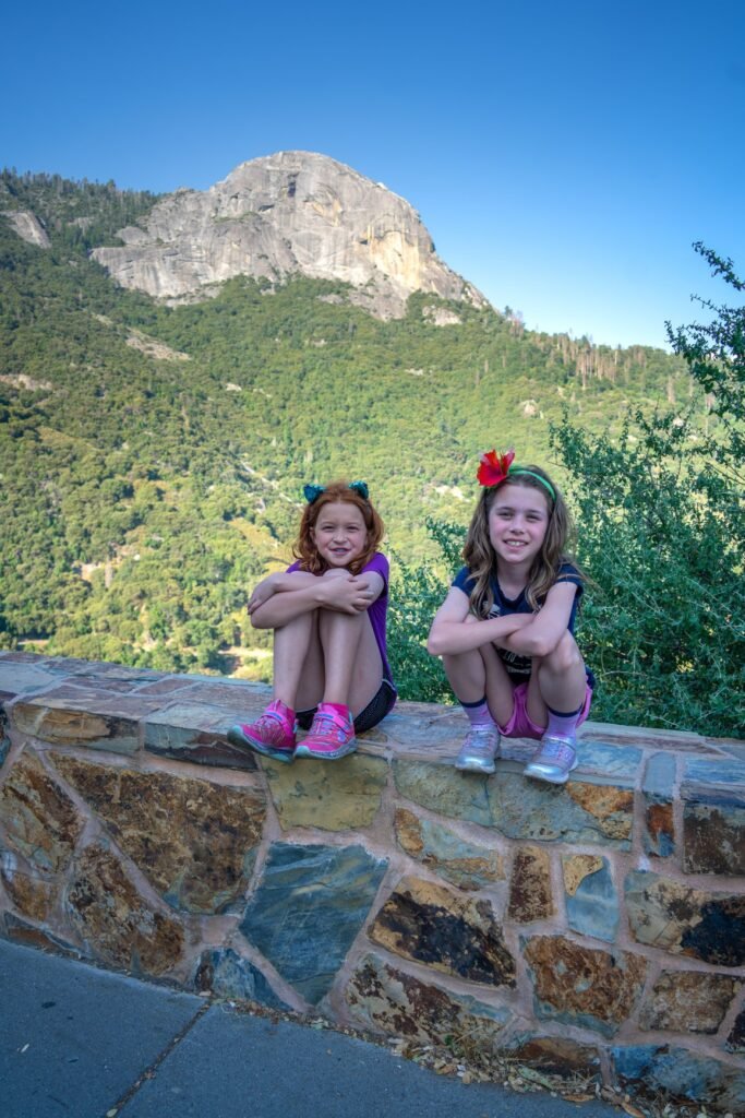 Two children sitting on a stone wall with a mountain and green forest in the background.