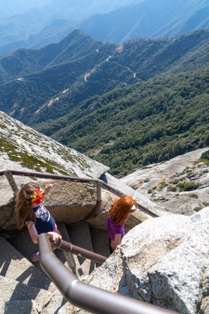 Two hikers descend a rocky mountain staircase with metal railings overlooking a vast green valley and distant peaks in the Pacific Northwest.