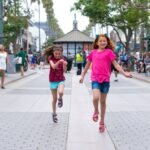 Two young individuals running happily along a tree-lined urban plaza with a gazebo and pedestrians in the background.