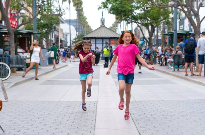 Two young individuals running happily along a tree-lined urban plaza with a gazebo and pedestrians in the background.