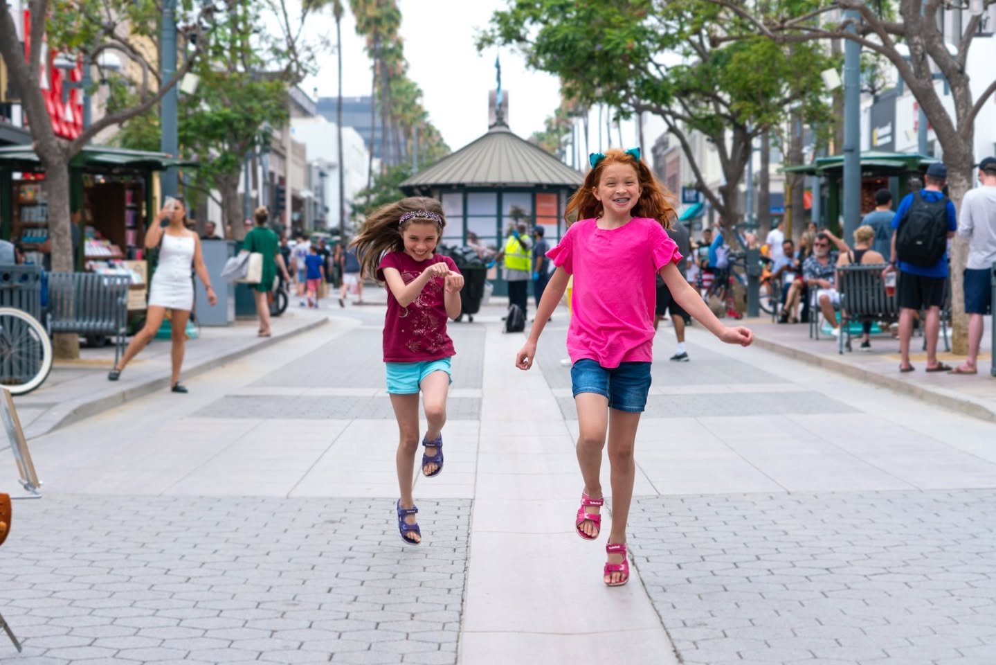 Santa Monica Two young individuals running happily along a tree-lined urban plaza with a gazebo and pedestrians in the background.