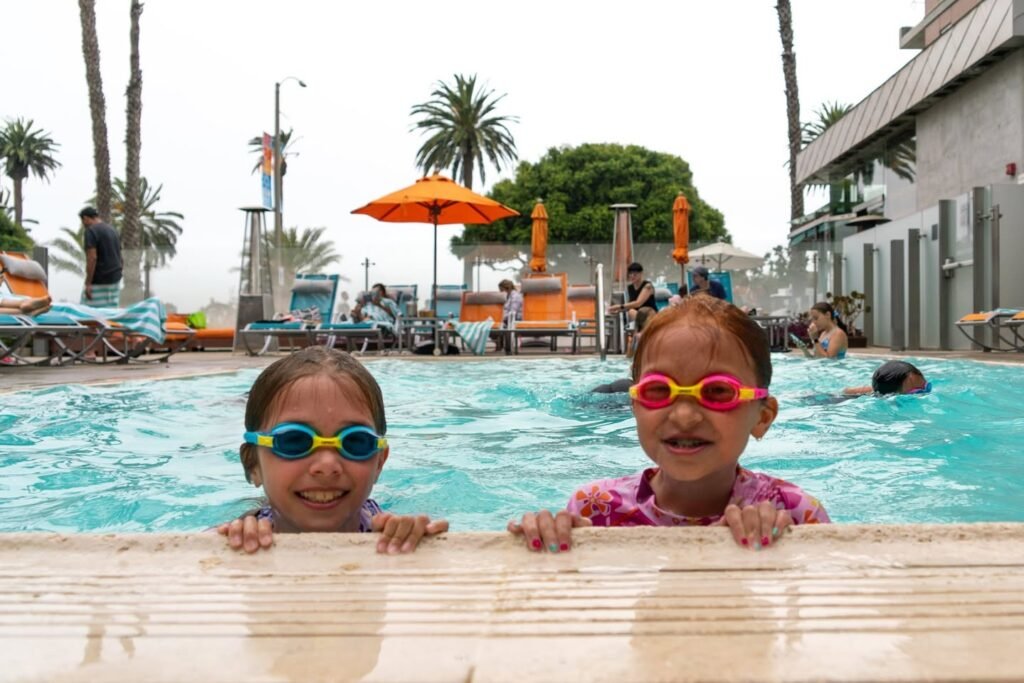 Children with colorful swimming goggles smiling at a resort pool. A view of the pool area with palm trees and loungers in the background.