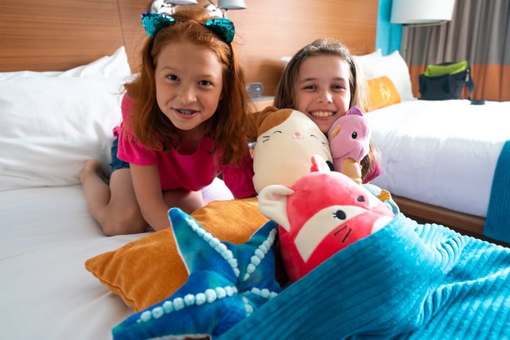 Two young girls smiling on a hotel bed surrounded by colorful plush toys and pillows.