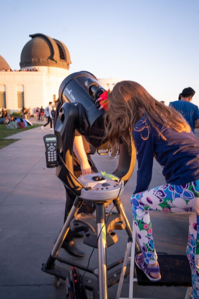 A person observing through a large telescope at a planetarium with domes in the background. Outdoor evening activity focused on astronomy equipment.
