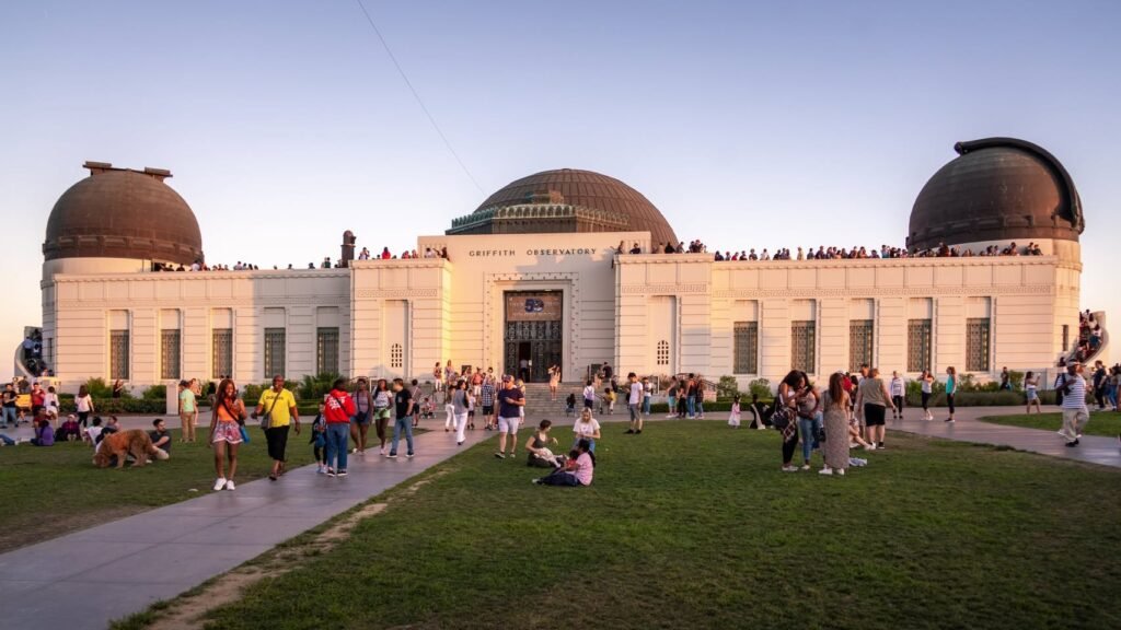 A colonial-style observatory building with domed roofs features crowds of visitors on surrounding grass and paved paths during evening. The site in Griffith Park, Los Angeles, is bathed in warm sunlight under a clear sky.