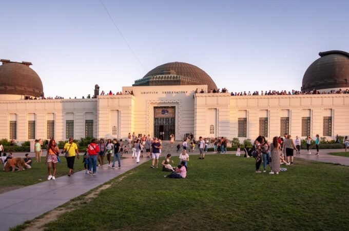 A colonial-style observatory building with domed roofs features crowds of visitors on surrounding grass and paved paths during evening. The site in Griffith Park, Los Angeles, is bathed in warm sunlight under a clear sky.