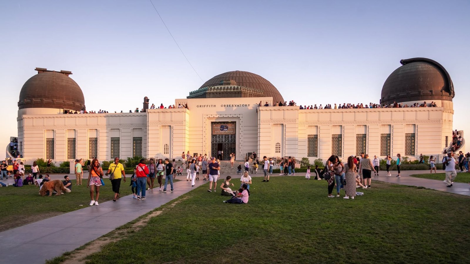 A colonial-style observatory building with domed roofs features crowds of visitors on surrounding grass and paved paths during evening. The site in Griffith Park, Los Angeles, is bathed in warm sunlight under a clear sky.