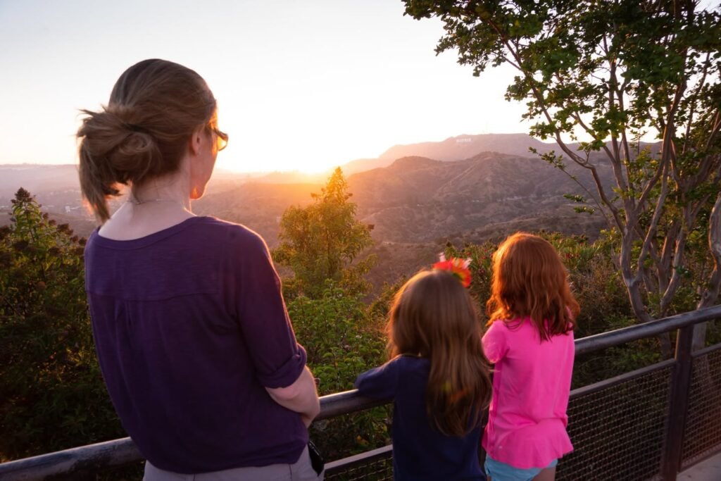 A person and children look out over rolling hills and mountains during a golden sunset. The scene includes trees and a railing on an observation deck.
