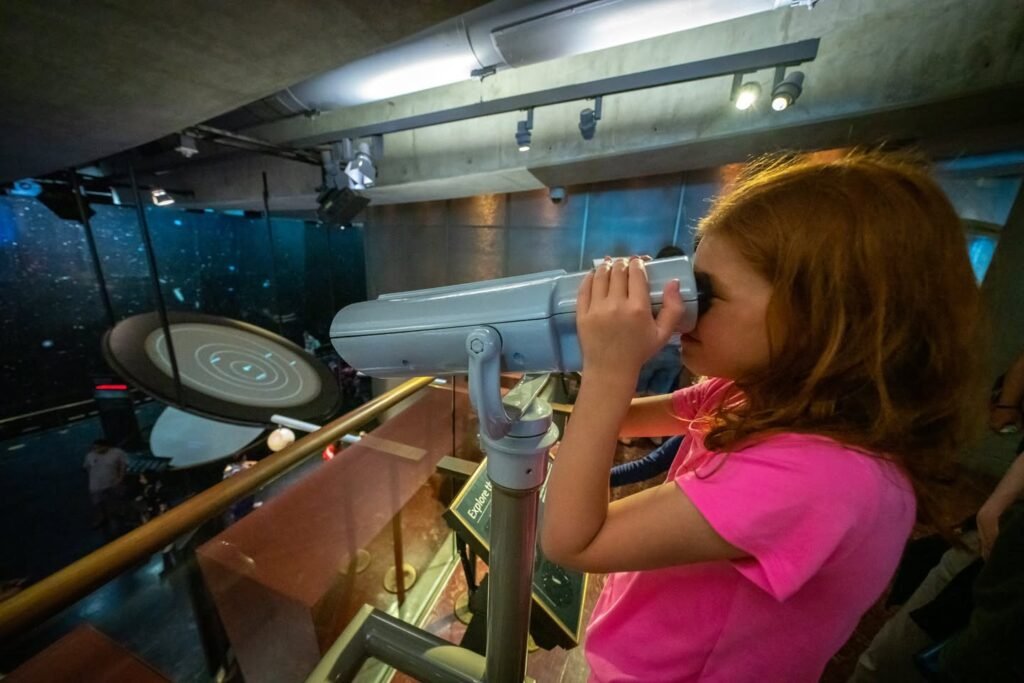 A child looks through a planetarium model equipped with interactive exhibits. The setting is an indoor science center, featuring educational models of the solar system.