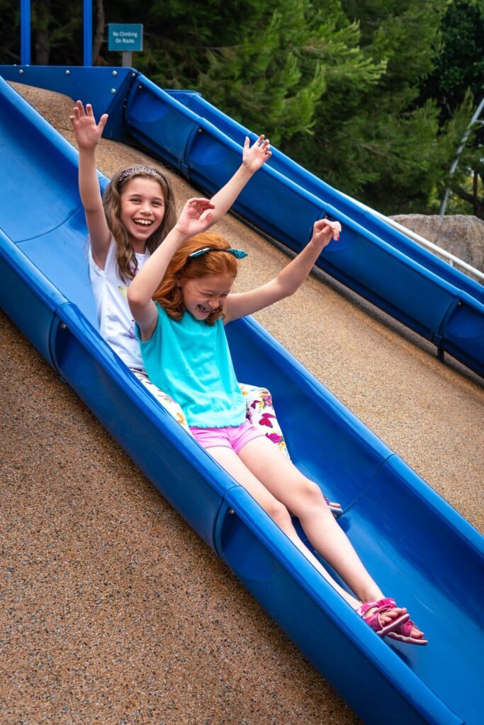 Children sliding down a blue playground slide with arms raised in excitement. Joyful moments captured at a park in the Pacific Northwest.