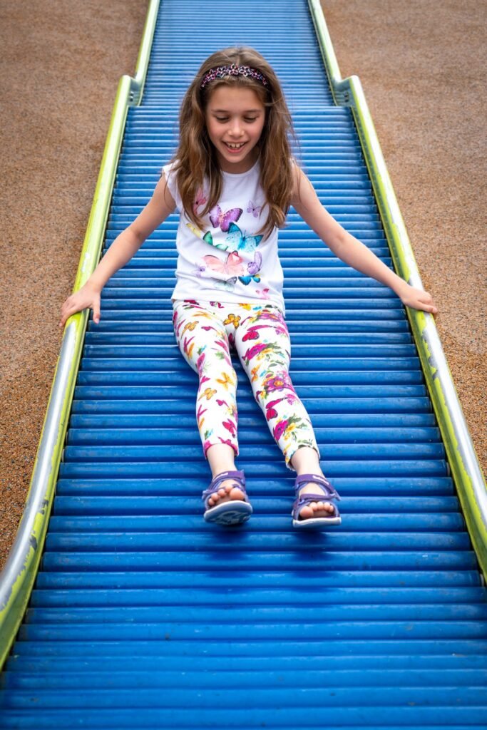 A happy child wearing floral leggings and a headband enjoys sliding down a bright blue corrugated playground slide.