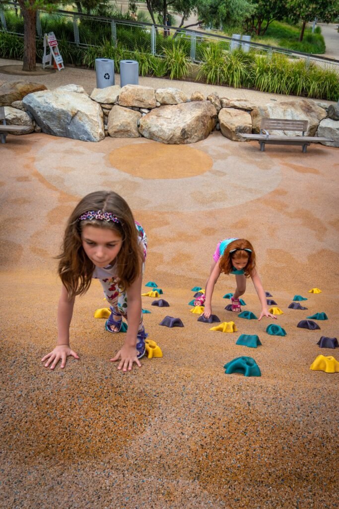 Two young girls scaling a colorful bouldering wall at the Summerlin Town Center climbing park. The playground features a large bouldering wall surrounded by rock formations and benches.