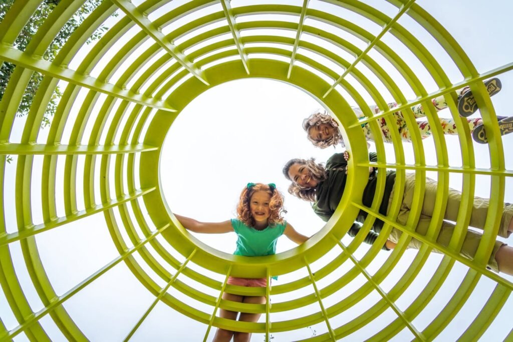 A smiling girl at the end of a bright yellow playground tunnel with adults entering behind her.