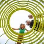 A smiling girl at the end of a bright yellow playground tunnel with adults entering behind her.