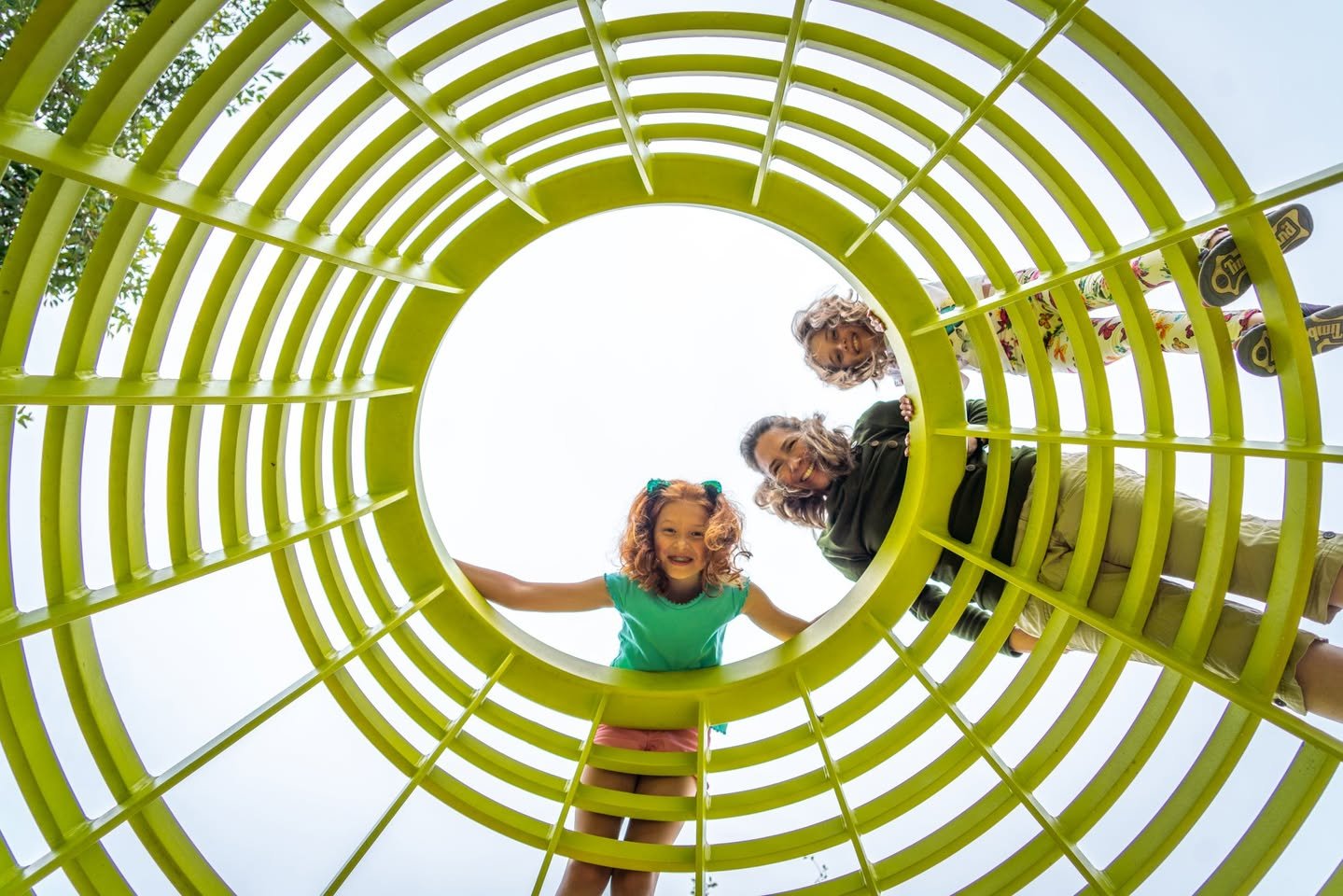 Tongva Park A smiling girl at the end of a bright yellow playground tunnel with adults entering behind her.