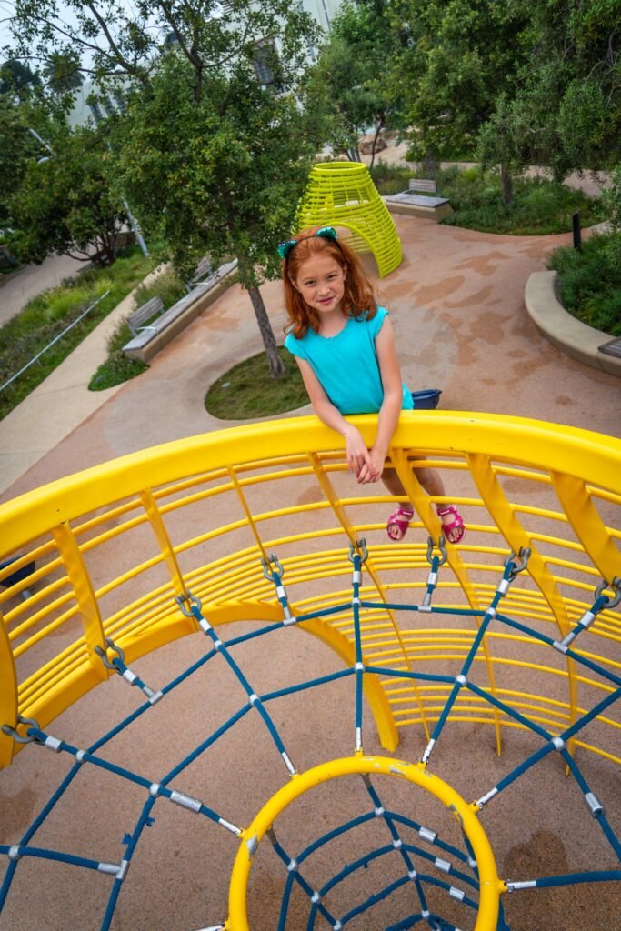 A young child sits on a yellow playground dome looking back at the camera in a green park setting.