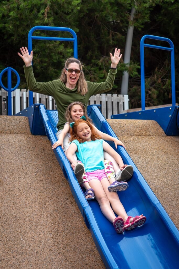 A woman and two children slide down a bright blue playground slide with joy, capturing a moment of shared family adventure.