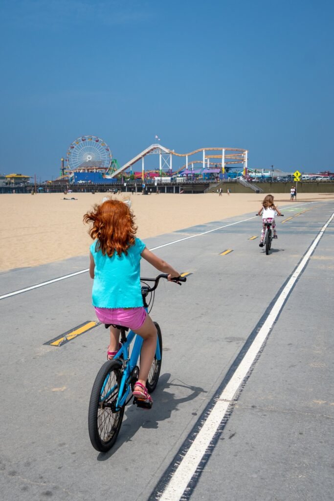 Two people riding bicycles on a paved path next to a sandy beach with amusement park rides in the distance under a clear blue sky.