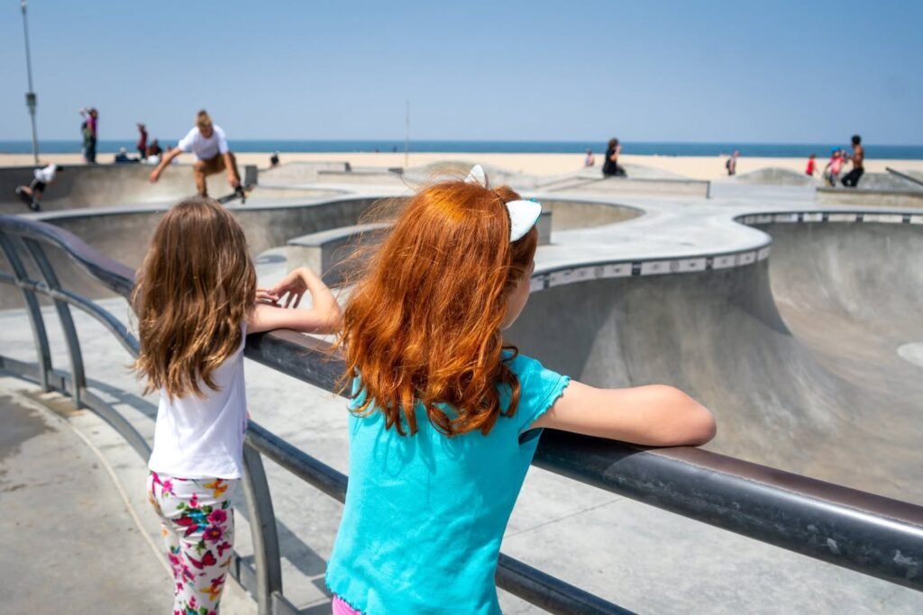 Two children watch skateboarders at a coastal skate park with the ocean in the background.