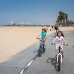 Tongva Park Children ride bicycles on a sunny day along a coastal path near Santa Monica beach, surrounded by palm trees and pedestrians.