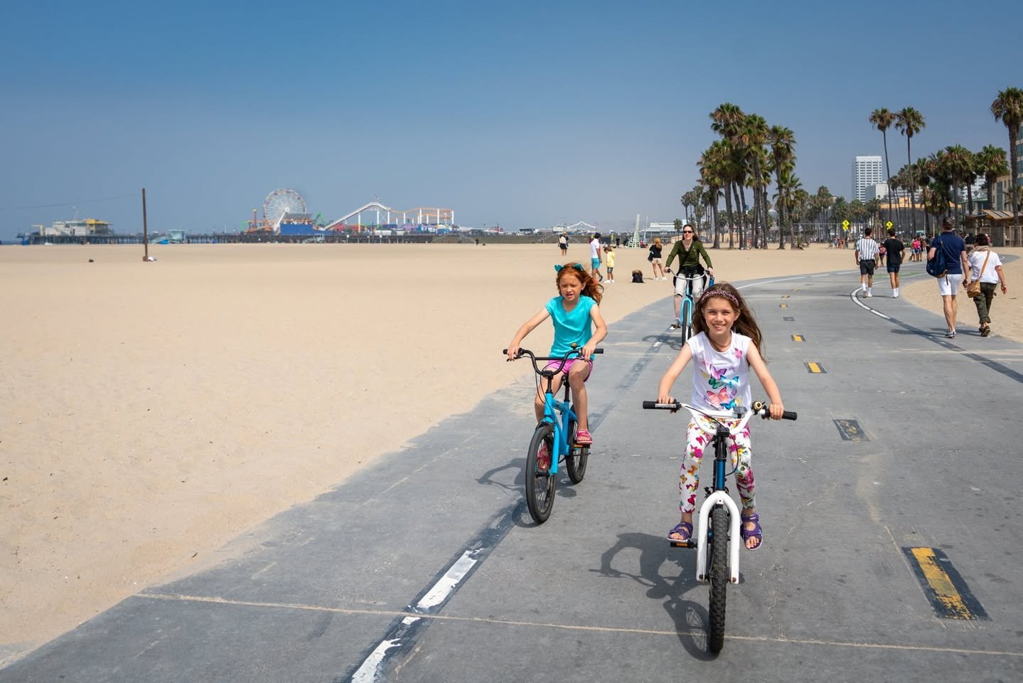 Pier to Pier Bike Ride Children ride bicycles on a sunny day along a coastal path near Santa Monica beach, surrounded by palm trees and pedestrians.