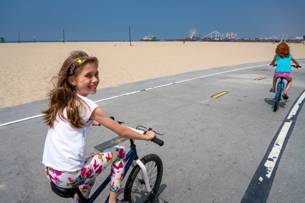 A young girl smiles while riding a bike on a paved path beside a sandy beach under a clear blue sky, with another cyclist ahead.