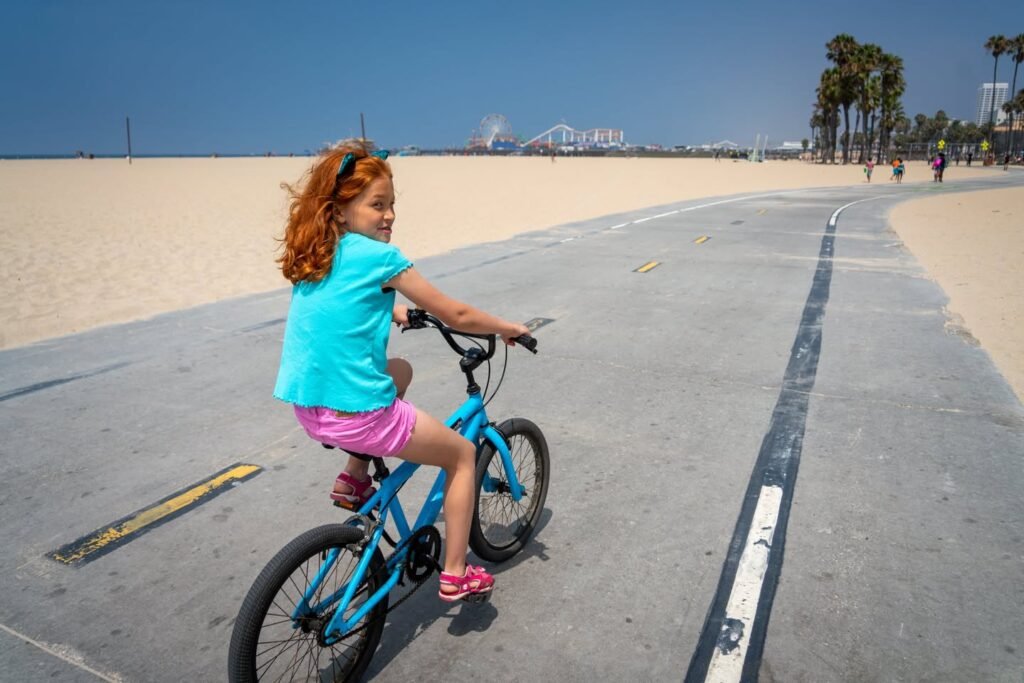 A child rides a blue bicycle along a paved beach path with sand and palm trees in the background.