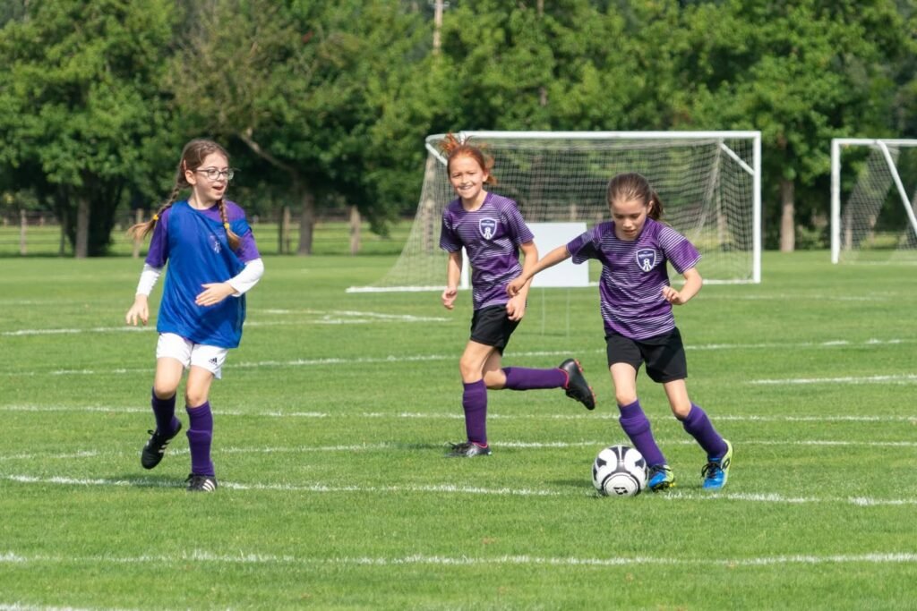 Young soccer competitors in purple and blue uniforms on a green field. White goal posts are visible in the background near a natural green tree line.